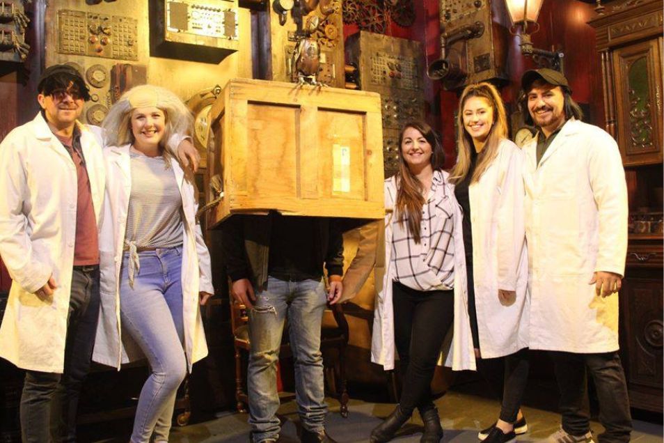 A group of people, some in lab coats, pose in an escape room with one person's head in a box.