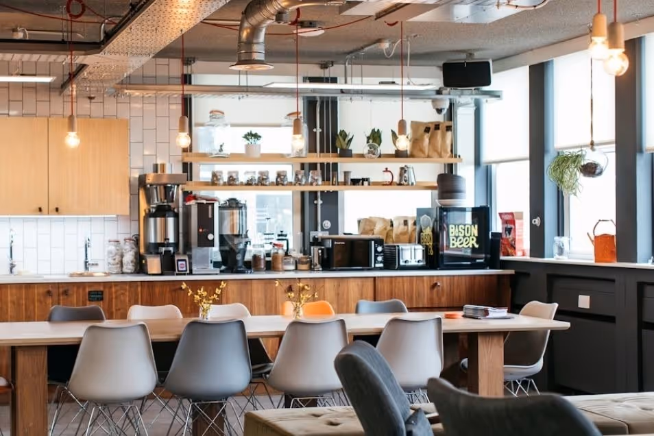 Office kitchen with a long wooden dining table, grey chairs, and hanging light fixtures.