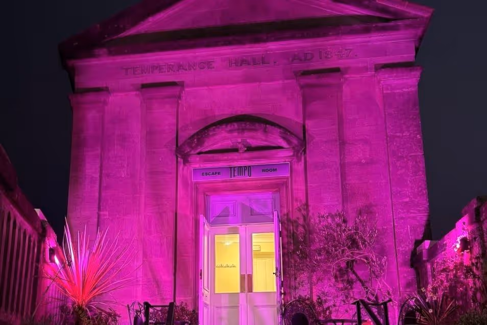 A stone building with an arched entrance is lit with purple lights at night.