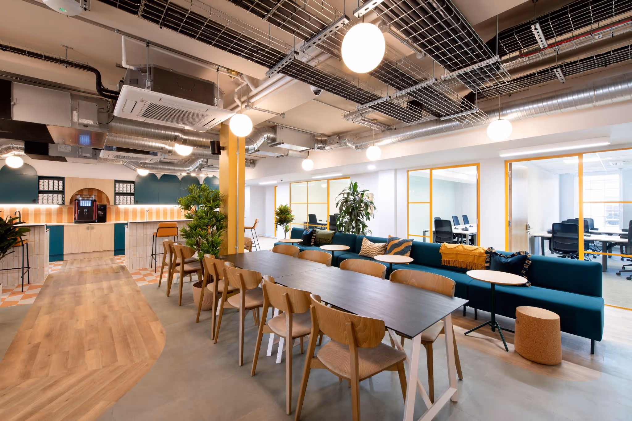 Coworking space featuring exposed industrial ceilings, a long black communal table with wooden chairs, and a deep teal velvet sofa.