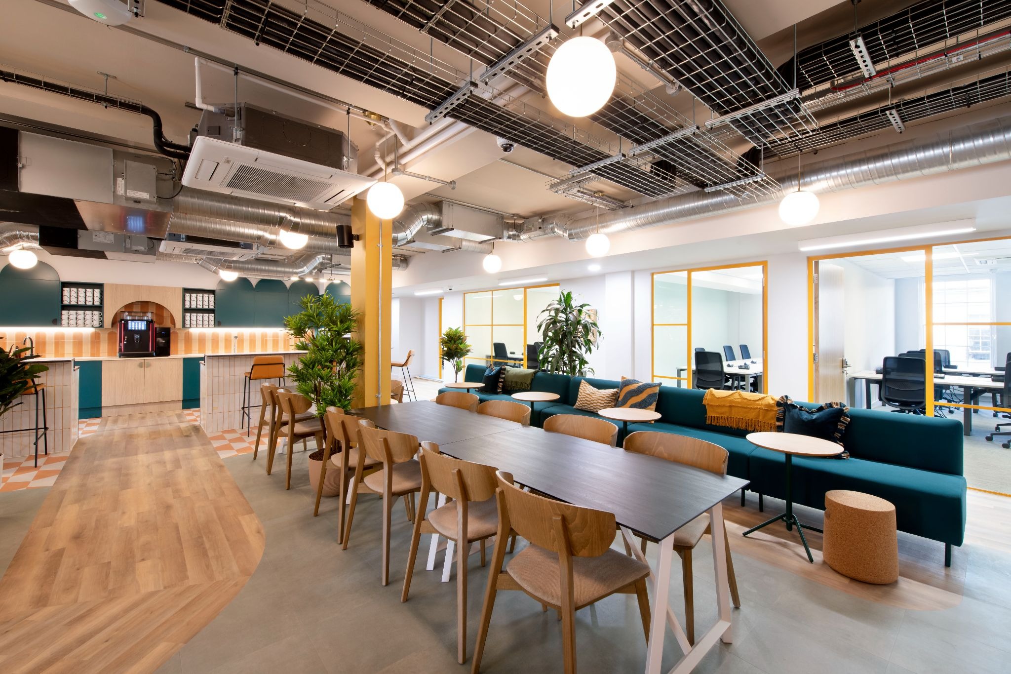 Coworking space featuring exposed industrial ceilings, a long black communal table with wooden chairs, and a deep teal velvet sofa.