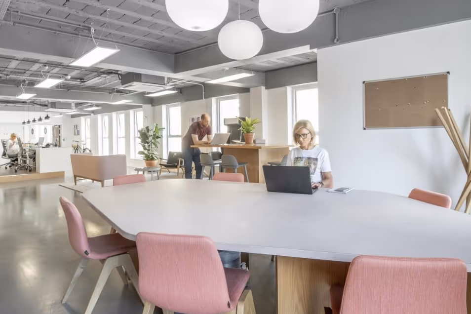 Bright, open-plan office featuring pink chairs at a large grey table, globe pendant lights, and people working in the space