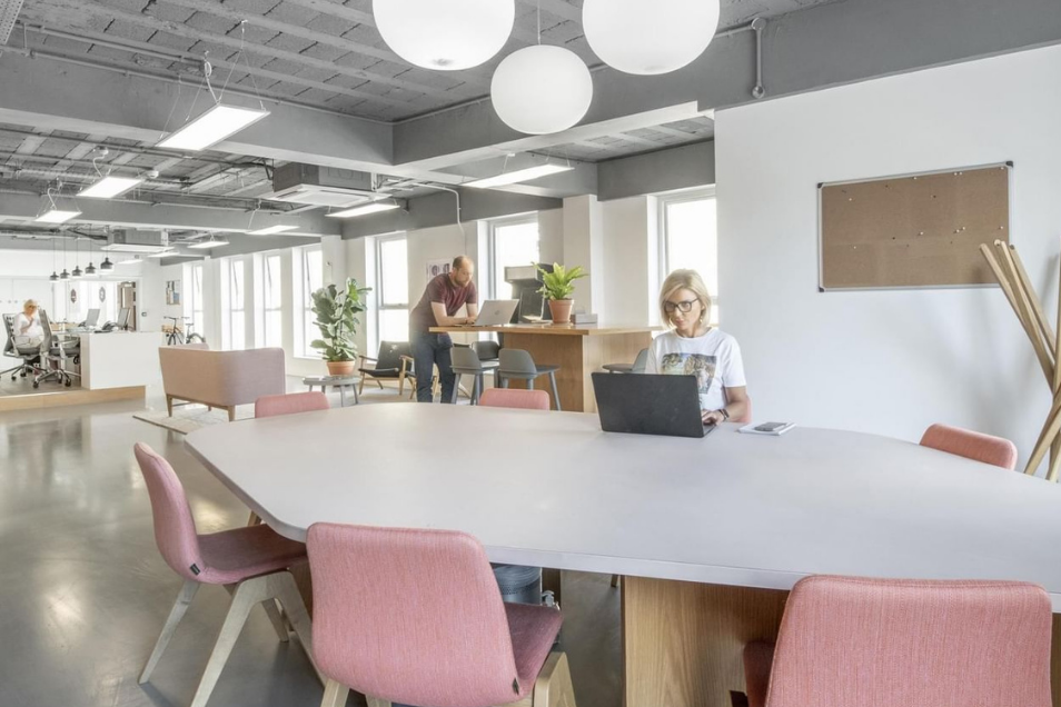 Bright, open-plan office featuring pink chairs at a large grey table, globe pendant lights, and people working in the space