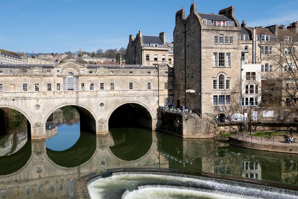 The historic Pulteney Bridge in Bath, with its arched stone structure and reflections in the river, features a weir in the foreground.