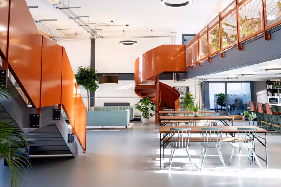 Office space featuring a prominent orange spiral staircase, wooden communal tables with grey chairs, and a bright mezzanine level.