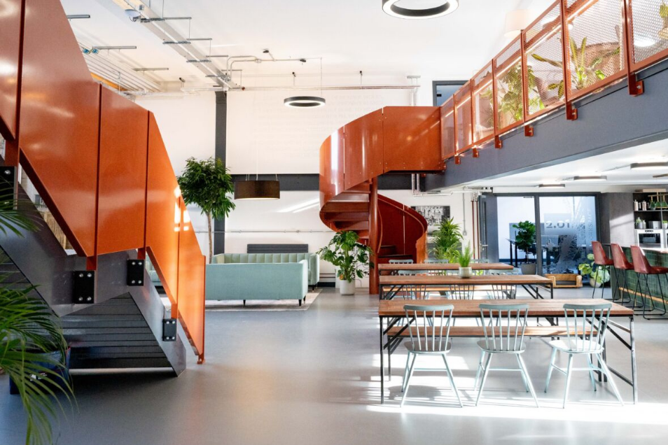 Office space featuring a prominent orange spiral staircase, wooden communal tables with grey chairs, and a bright mezzanine level.