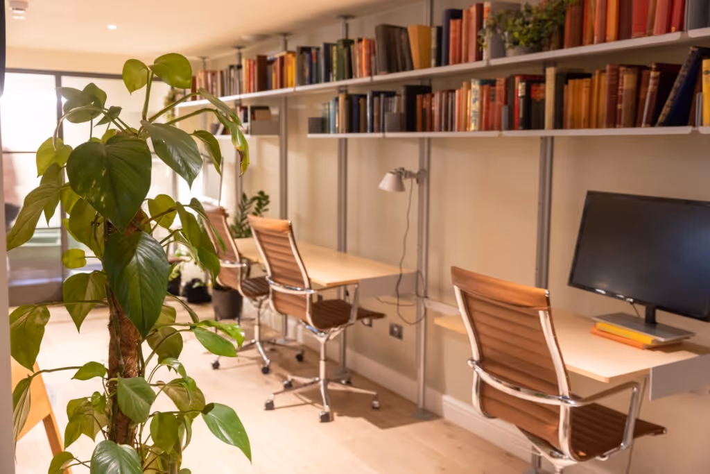 Cosy home office featuring a wall-mounted shelving unit with books, wooden desks, ergonomic chairs, and a large leafy plant.