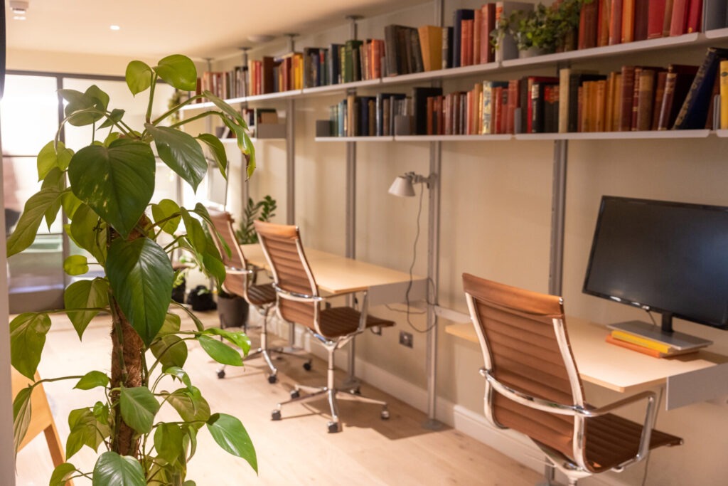 Cosy home office featuring a wall-mounted shelving unit with books, wooden desks, ergonomic chairs, and a large leafy plant.