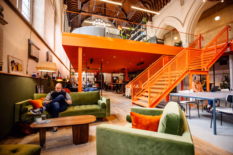 Large open-plan office in a repurposed church, featuring a prominent orange mezzanine and staircase above olive green velvet lounge seating.