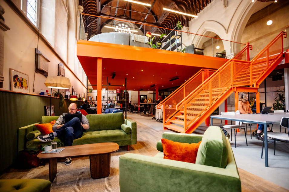 Large open-plan office in a repurposed church, featuring a prominent orange mezzanine and staircase above olive green velvet lounge seating.