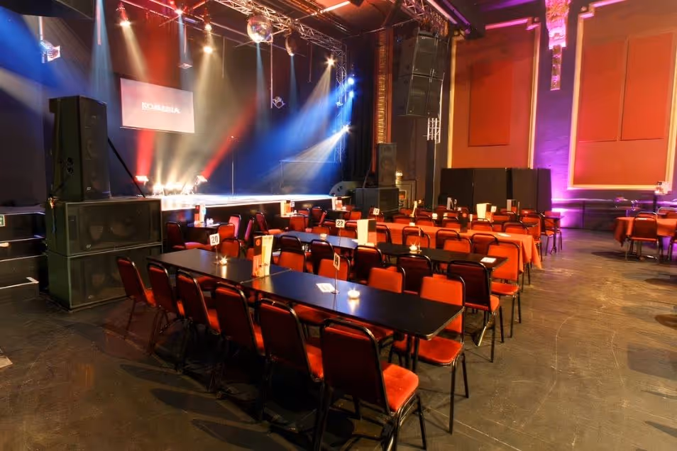 A large event space or concert hall with long tables and red chairs arranged in front of a stage with theatrical lighting.