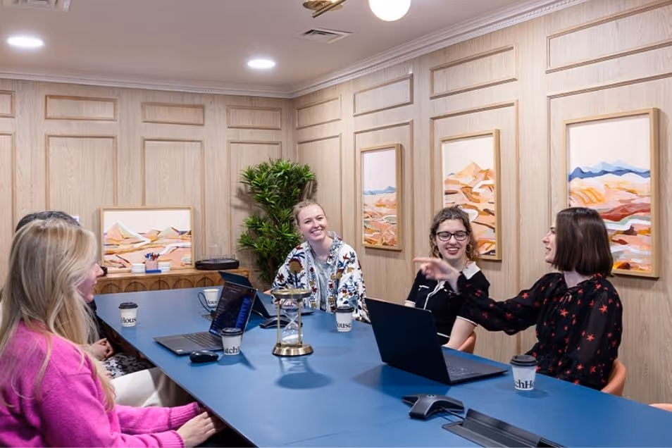 Four women sat at a large blue conference table, smiling and conversing during a business meeting in a panelled room.