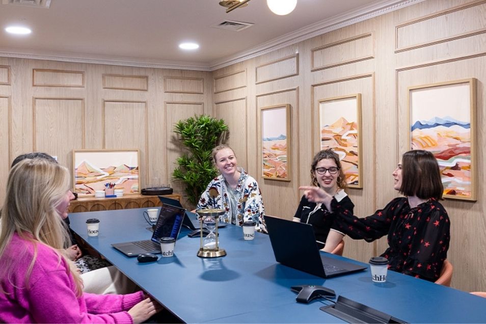 Four women sat at a large blue conference table, smiling and conversing during a business meeting in a panelled room.