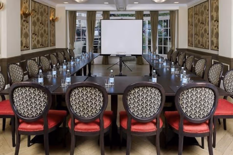 A formal meeting room set up with a U-shaped table, patterned chairs, and a white presentation screen.