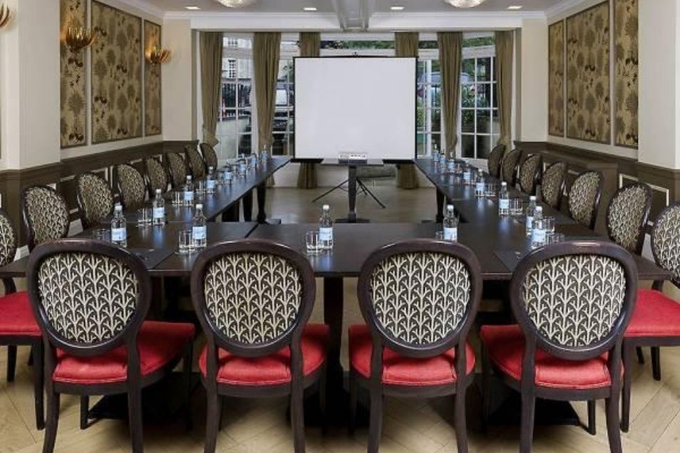 A formal meeting room set up with a U-shaped table, patterned chairs, and a white presentation screen.
