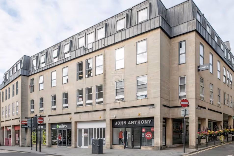 Modern multi-story stone building with ground-floor shops, including "ROSS ANTHONY," and a grey roof with dormer windows.