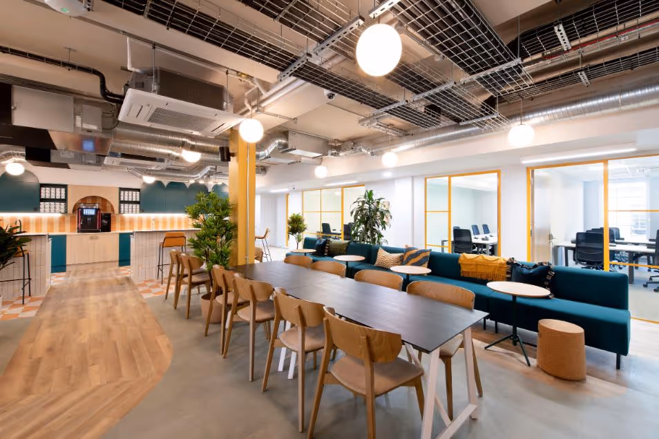 Office lounge with an exposed ceiling, featuring a long dark wood table and a deep blue velvet sofa with yellow accents.