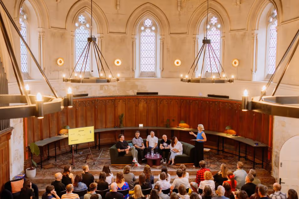 A panel discussion taking place in a circular, historic hall with arched windows, large chandeliers, and an audience seated in the foreground.