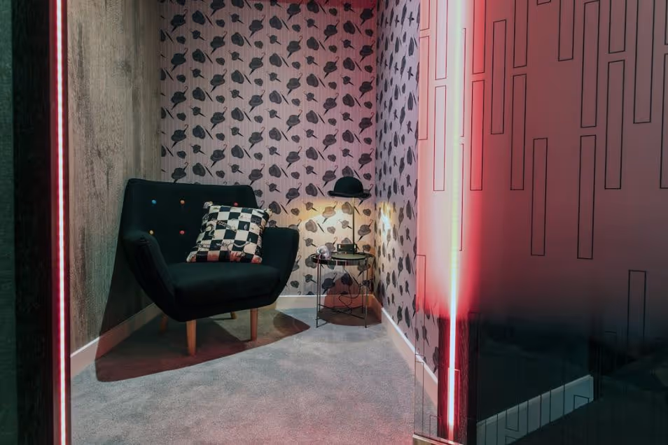 Lounge nook with patterned floral wallpaper, a black armchair with colorful buttons, and a glowing neon pink light fixture.
