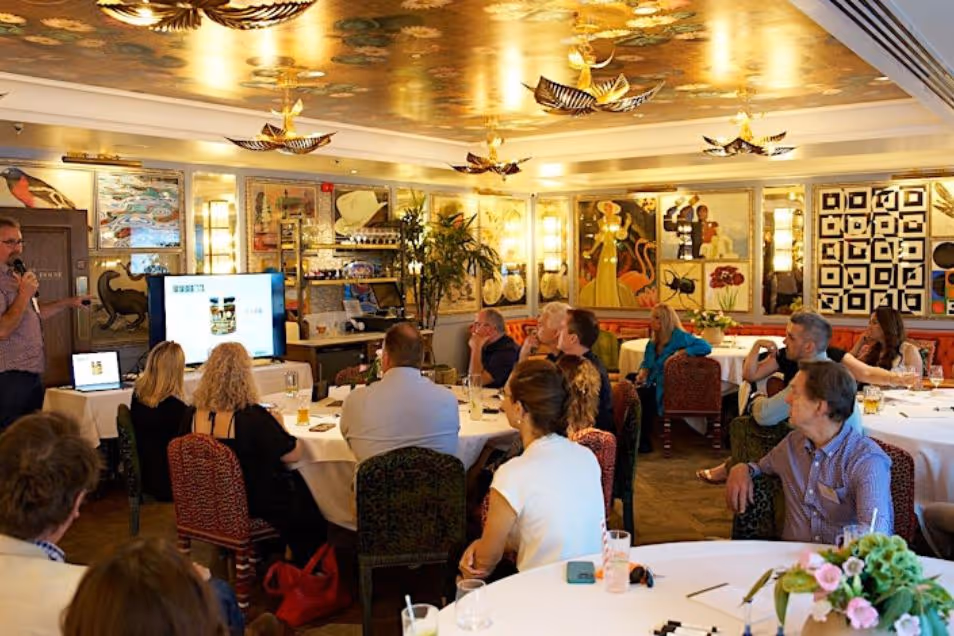 People sit at round tables in a dining room while a person stands and speaks to the group.