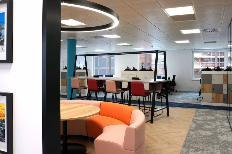 Bright office interior showing a pink and orange curved sofa, high-top collaborative desks, and wooden storage lockers.