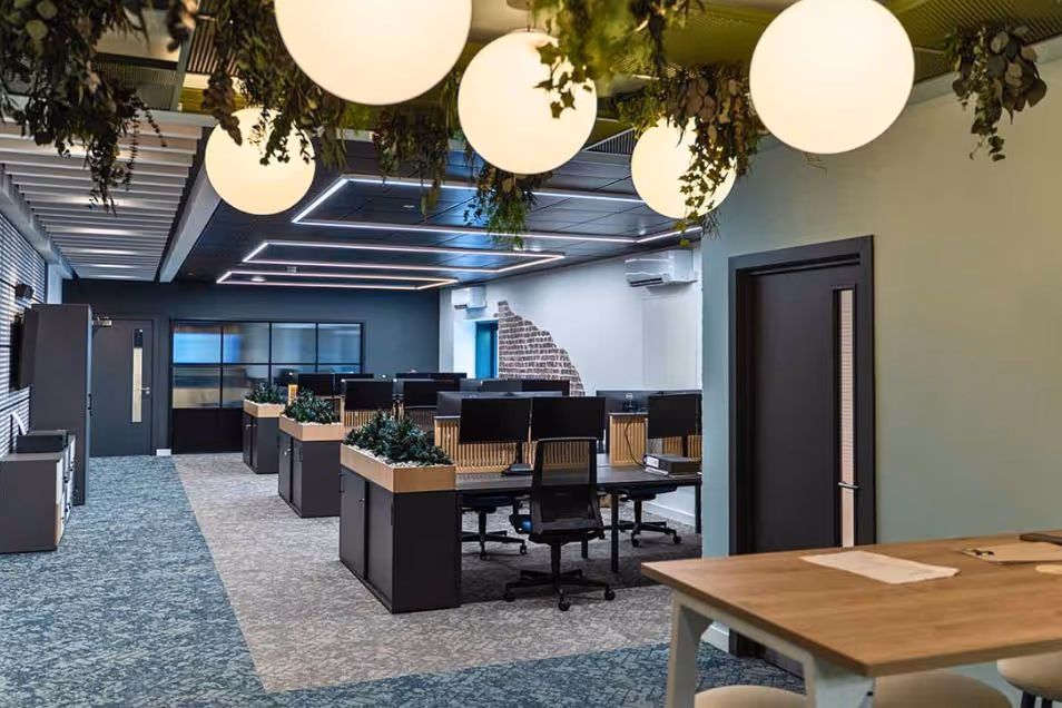 Open-plan workspace featuring black desks, blue carpeting, and large globe pendant lights draped with hanging greenery.