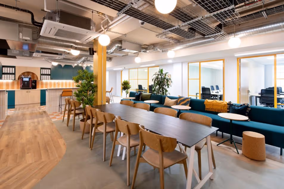 Office break room with a long dark communal table, wooden chairs, and a deep teal sofa under industrial lighting.