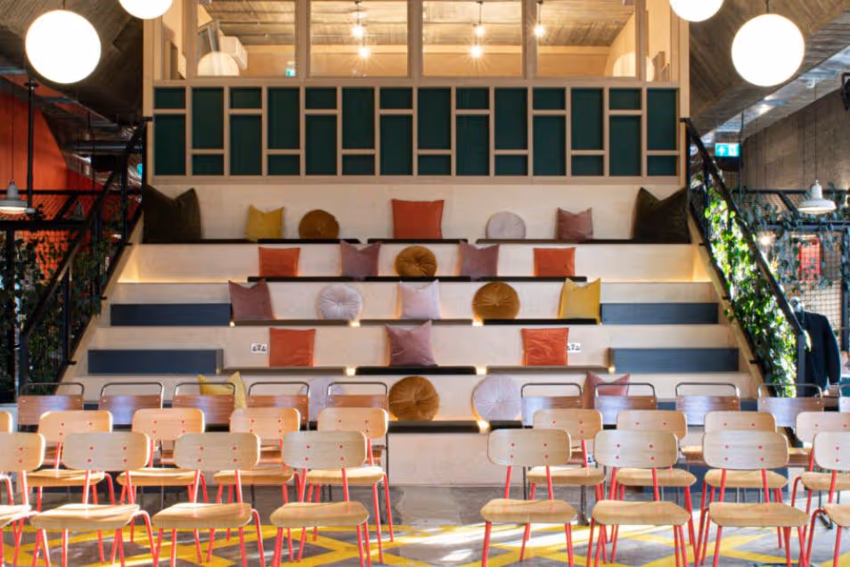 Rows of chairs facing tiered wooden seating with cushions in an event space under pendant lights.