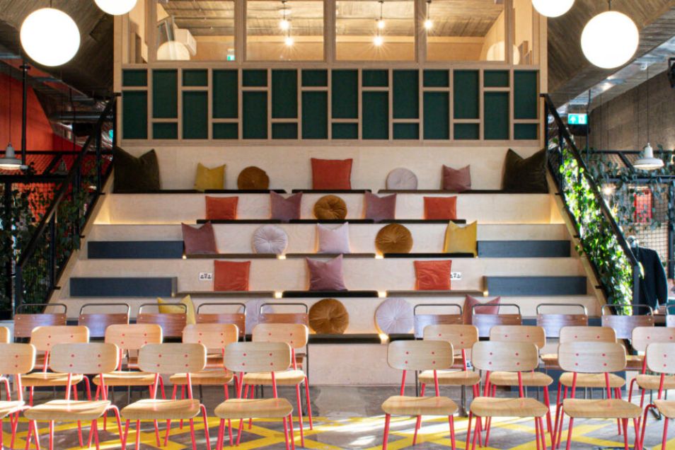 Rows of chairs facing tiered wooden seating with cushions in an event space under pendant lights.