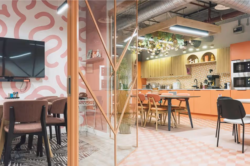 A view through a glass partition of an office kitchen with tables, chairs, and a pink tiled floor.
