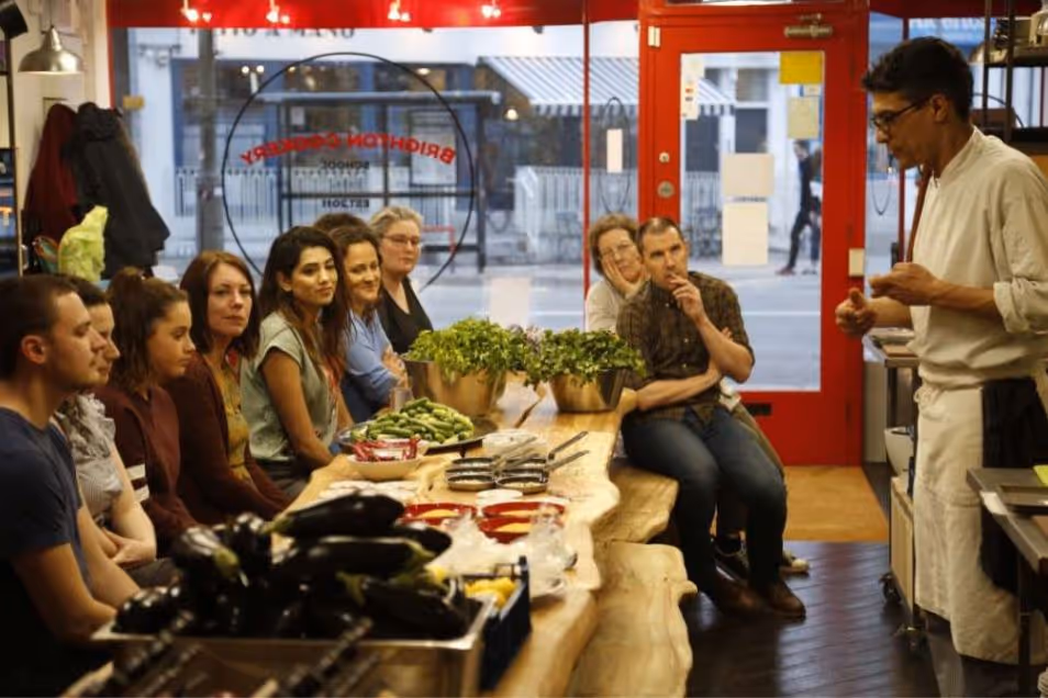 A chef speaks to a group of people sitting at a wooden table during a cooking class.