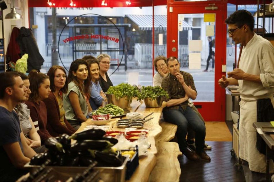 A chef speaks to a group of people sitting at a wooden table during a cooking class.