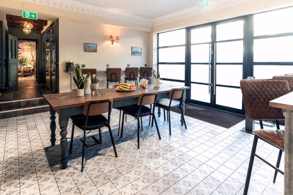 A long wooden table with black chairs on a tiled floor in a room with large glass doors.