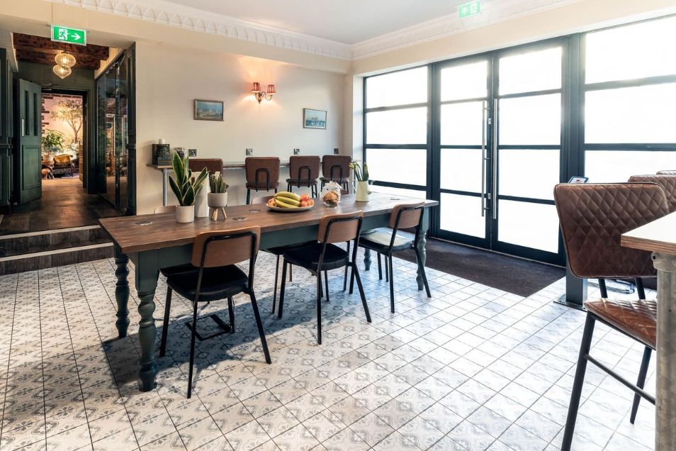 A long wooden table with black chairs on a tiled floor in a room with large glass doors.