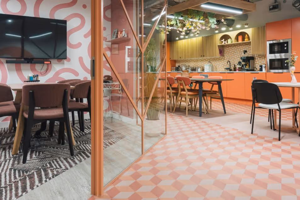 An office kitchen and dining area with orange cabinets, a patterned floor, and a meeting room behind a glass wall.