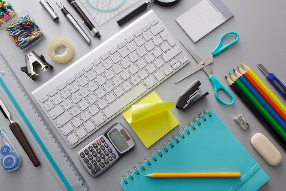 Top-down view of a desk with keyboard, calculator, sticky notes, pens, scissors and neatly arranged stationery on a grey surface.