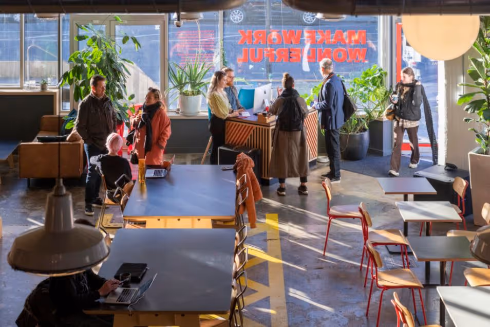 A busy office cafe where people stand and chat near a counter and sit at black tables in a sunlit space.