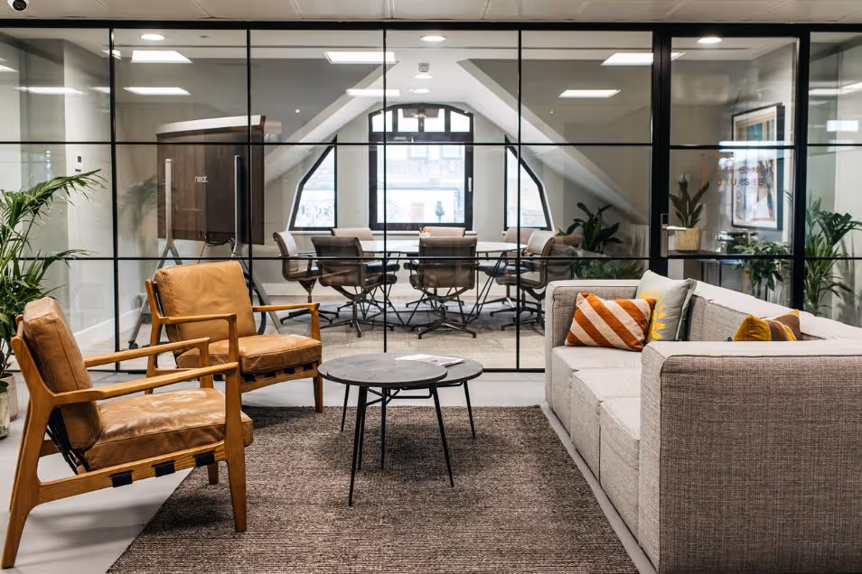 Office lounge with tan leather armchairs and a grey sofa, separated from a meeting room by a glass wall.