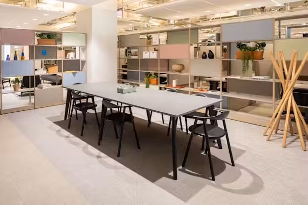A grey table with black chairs sits in an office near a wall of shelves and a wooden coat rack.