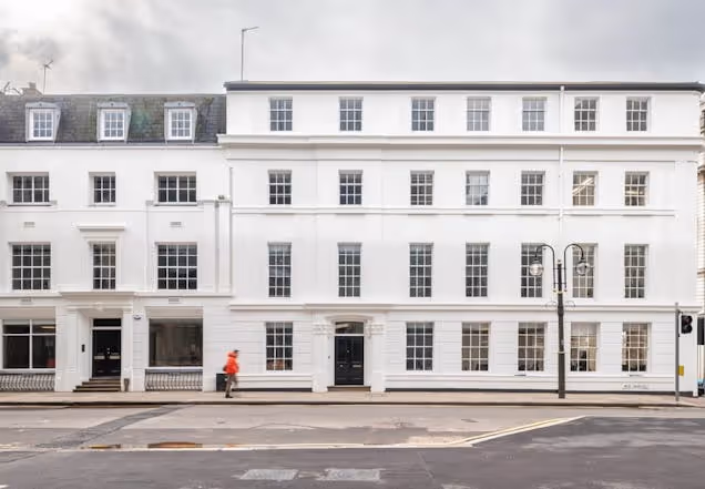 A person walks past a white four-story building with many windows and black doors on a city street.