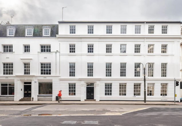 A person walks past a white four-story building with many windows and black doors on a city street.