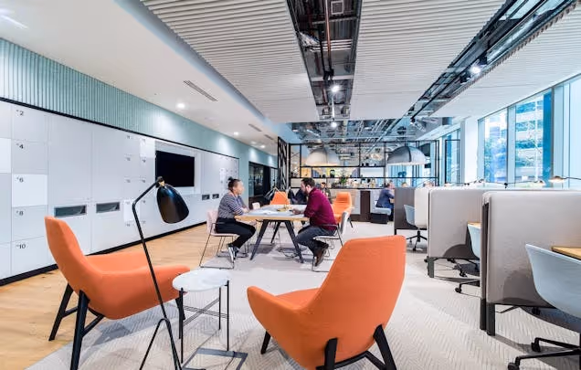 People sat at tables and in orange armchairs while working in a long office with a textured ceiling.