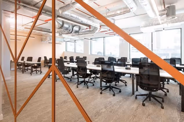 Rows of white desks with black chairs in an office featuring orange metal beams and exposed ceiling pipes.