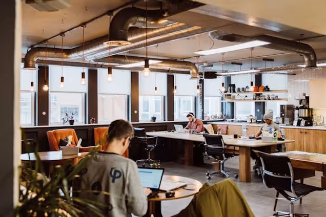 People sitting at wooden tables working on laptops in an office with industrial ceiling pipes and windows.