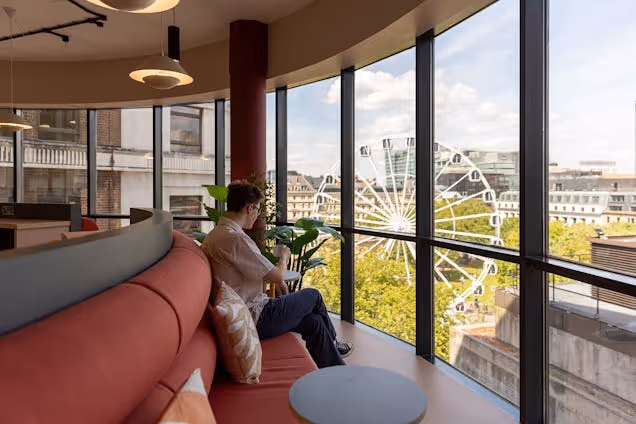 A person sitting on a red sofa in a breakout space looking out a window at a city with a Ferris wheel.