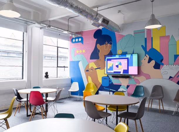 A room with tables, chairs, and a mural on the wall featuring faces and shapes next to windows.