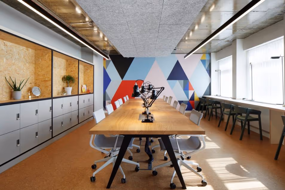 A wooden table with lamps and black chairs in a coworking space with a geometric mural on the wall.