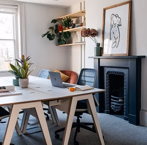 A laptop on a trestle table in an office with a fireplace, plants, and shelving.