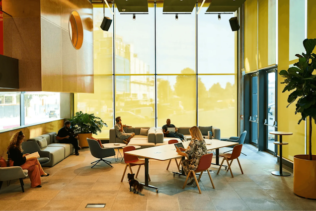People at tables and on sofas in a lounge area with floor-to-ceiling windows and plants.