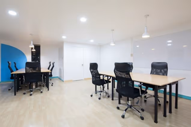 Black chairs at wooden tables in a room with a whiteboard and spotlights on the ceiling.